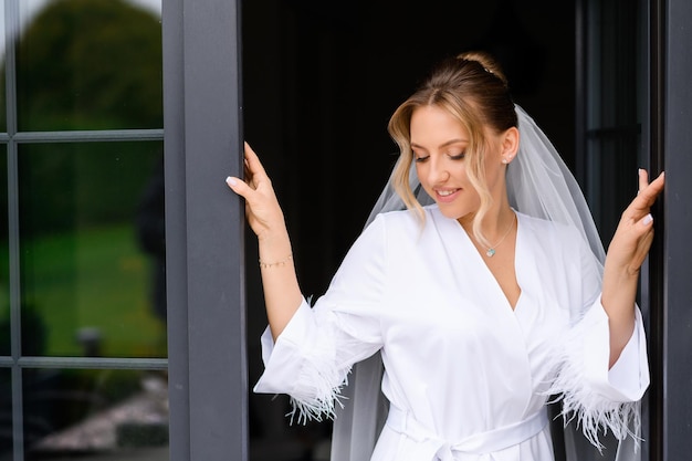 Free photo close view of attractive bride with blonde hair and natural makeup wearing in white and silk peignoir looking down and posing near opened door on balcony