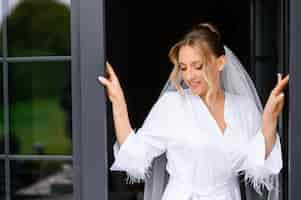 Free photo close view of attractive bride with blonde hair and natural makeup wearing in white and silk peignoir looking down and posing near opened door on balcony