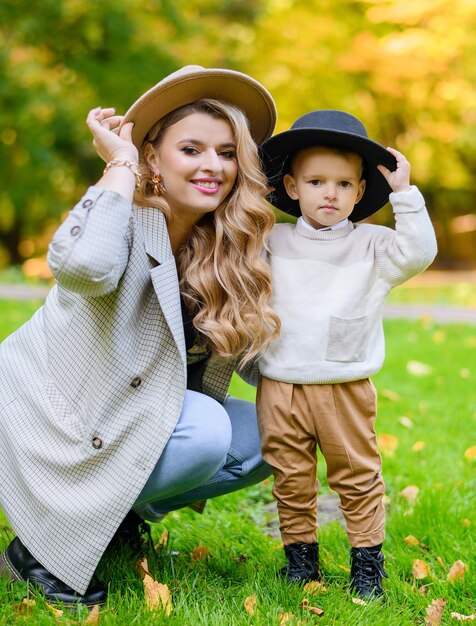 Close view of attractive woman and boy in casual clothes sitting and holding hats on heads smiling and posing at camera during sunny autumn day in park