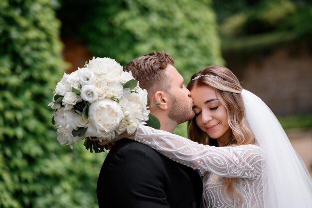 Close view of bearded groom in black suit kissing bride which hugging his neck with flowers and closing eyes while posing on nature during wedding event