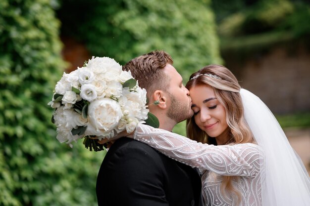 結婚式のイベント中に自然にポーズをとっている間、花で首を抱き締めて目を閉じている花嫁にキスをしている黒いスーツを着たひげを生やした新郎の拡大図