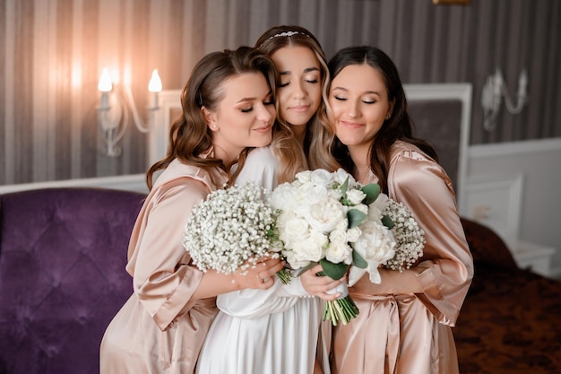 Close view of beautiful bridesmaids in pastel peignoirs standing with both sides of bride and hugging her closing eyes and holding amazing bouquets of flowers