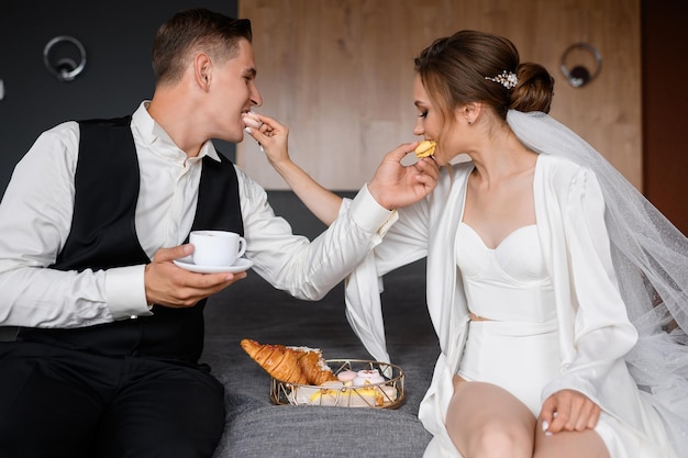 Close view of beautiful couple in trendy clothes sitting together on bed near plate with croissants and cookies feeding each other macarons and spending wedding morning together in hotel room