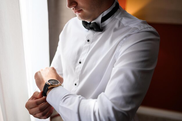 이른 아침에 결혼식을 준비하는 동안 왼쪽 손목에 값비싼 시계를 차고 있는 세련된 흰색 셔츠와 검은색 나비 넥타이를 한 얼굴 없는 신랑의 클로즈업