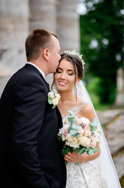 Close view of handsome bridegroom in black suit standing and kissing wife face while she feeling happy and hugging his during wedding walk outdoor