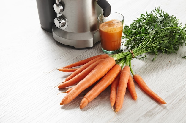 Close view metallic professional juicer with glass filled with tasty juice for breakfast from organic farm carrots lying on wooden table.