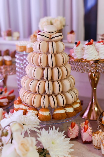 Close view of set of pastel macaroons which arranged in form of pyramid on glass plate and surrounded by tasty desserts with fruits and golden trails with cakes which placed on candy table