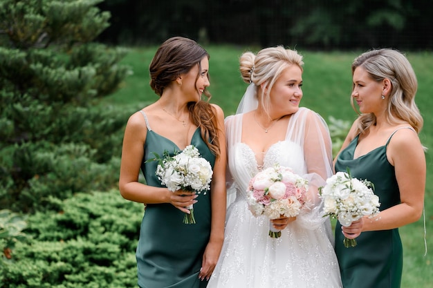 Близкий вид потрясающих подружек невесты в элегантных шелковых платьях с букетами цветов, позирующих с невестой на природе в день свадьбы