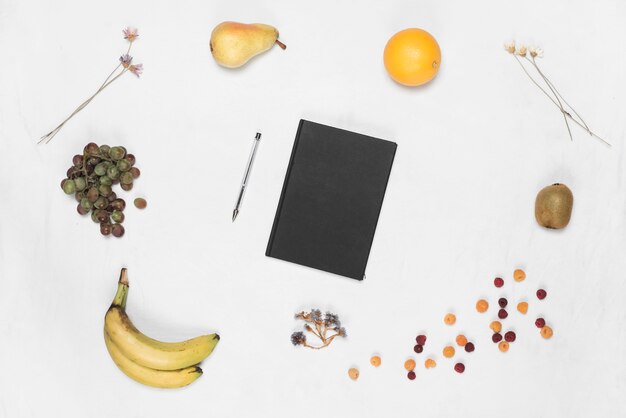 Closed black cover diary and pen with many fruits on white backdrop