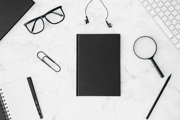 Closed black diary with eyeglasses; earphone; magnifying glasses; pencil; keyboard and diary on white desk