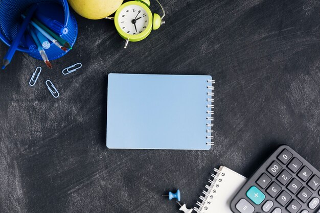 Closed blue notepad surrounded with stationery on chalkboard