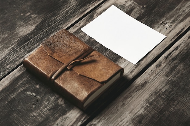 Закрытый блокнот в обложке из натуральной кожи рядом с листом белой бумаги на черном винтажном матовом деревянном столе фермы