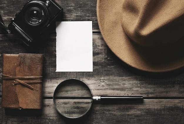 Closed notebook in leather cover, sheet of white paper, felt brown hat, camera and big magnifier isolated on black aged wood table