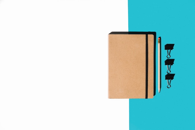 Closed notebook with brown cover; pencil and bulldog clips on white and blue background