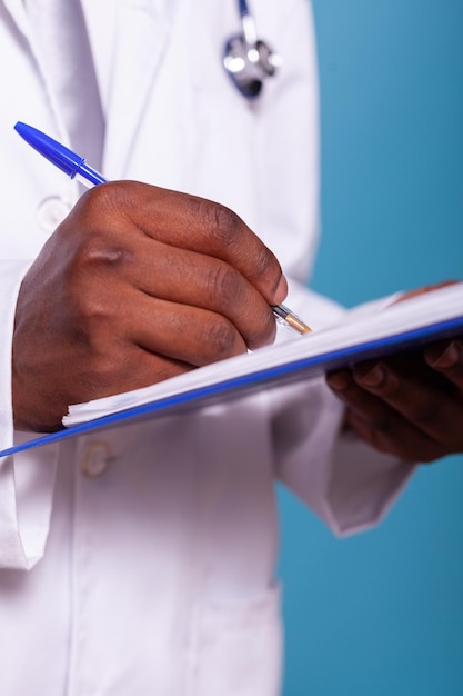 Крупный план афро-американского врача, держащего шариковую ручку в буфере обмена с медицинской картой пациента. Обрезанный подробный вид медика, делающего заметки на диаграммах результатов тестов.