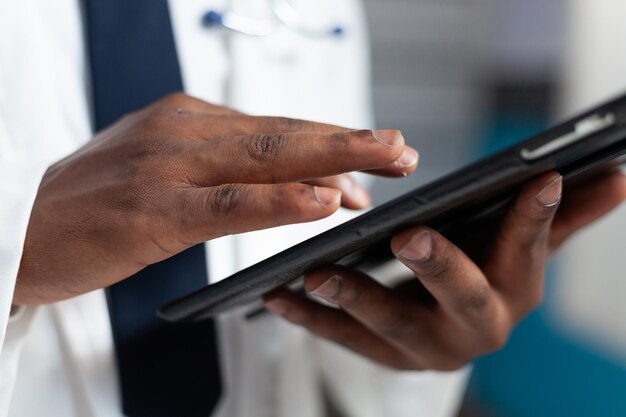 Closeup of african american doctor holding tablet computer typing healthcare treatment after analyzing disease symptoms during clinical appointment. Therapist man working in hospital office