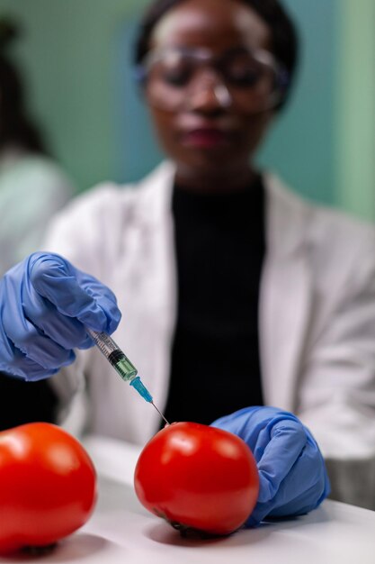 微生物学実験中に農薬を有機トマトに注入する医療用手袋を着用したアフリカ系アメリカ人の研究者のクローズアップ。 gmo野菜をテストする農業病院の実験室で働く生化学者