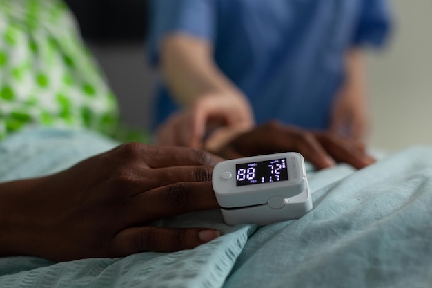 병원 병동에서 임상 상담을 하는 동안 손가락에 의료 산소 측정기를 들고 침대에서 쉬고 있는 아프리카계 미국인 환자의 클로즈업. 심장 박동수를 모니터링하는 여성 간호사. 의료 서비스