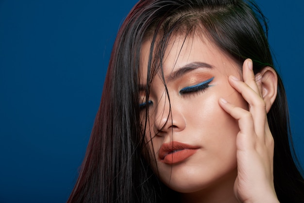 Closeup of Asian woman with full makeup posing in studio with closed eyes and fingers on temple