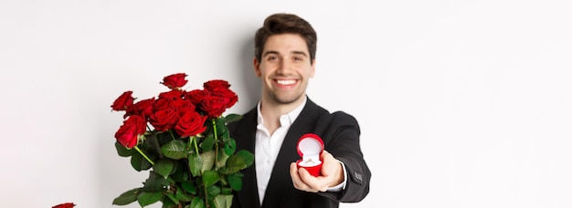 Primo piano dell'uomo attraente in vestito che tiene il mazzo delle rose che dà un anello di fidanzamento che propone a gi