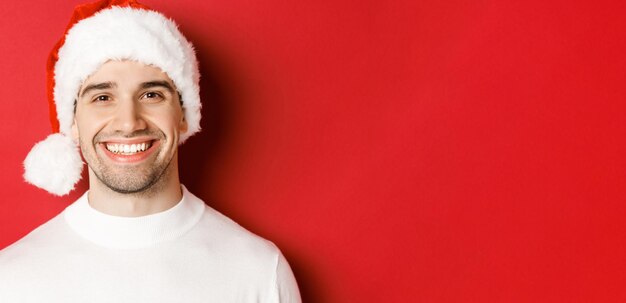 흰색 스웨터와 산타 모자를 쓴 매력적인 웃는 남자가 빨간색 배경에 서서 겨울 휴가를 즐기고 있는 모습을 클로즈업