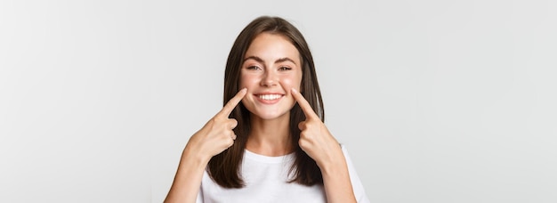 Closeup of attractive smiling young girl pointing fingers at face poking cheeks white background