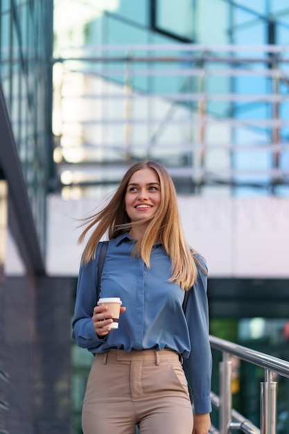 Closeup attractive woman in motion with takeaway coffee on business building. Portrait blonde girl holding paper cup with hot drink outdoor.
