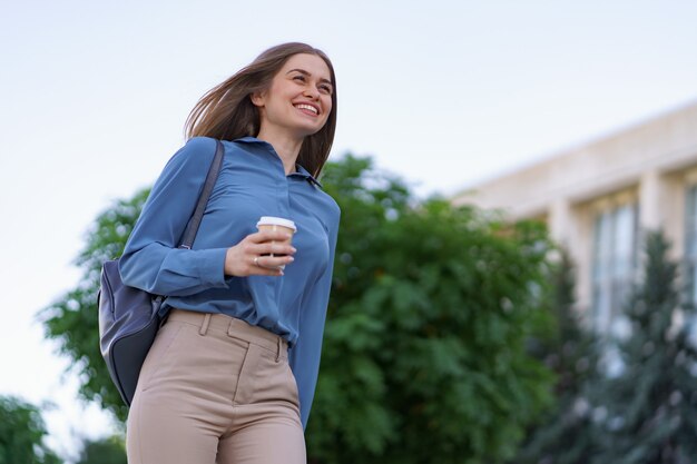 街でテイクアウトのコーヒーと動きのある魅力的な女性をクローズアップ。屋外の温かい飲み物と紙コップを保持している肖像画のブロンドの女の子。