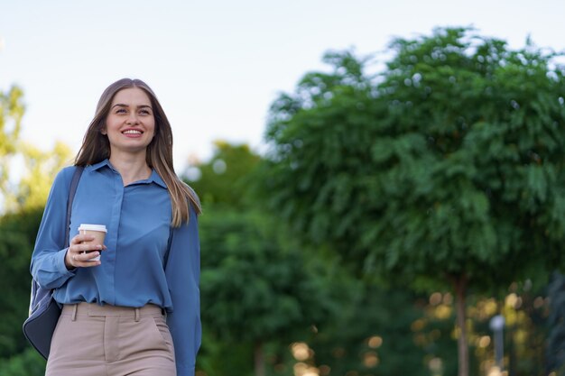 街でテイクアウトのコーヒーと動きのある魅力的な女性をクローズアップ。屋外の温かい飲み物と紙コップを保持している肖像画のブロンドの女の子。