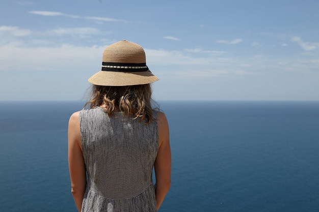 Closeup back view of unrecognizable slender blonde woman in dress and straw hat enjoying amazing seascape at viewpoint. romantic female admiring picturesque scenery over vast calm ocean and blue sky