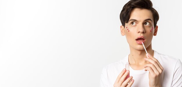 Closeup of beautiful androgynous man with glitter under eyes applying lip gloss looking dreamy at upper left corner white background