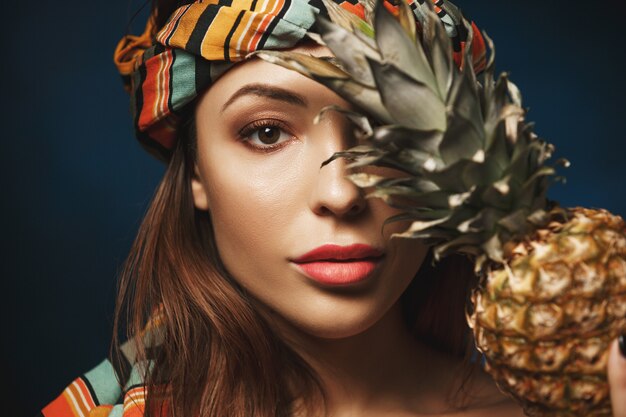 Closeup of beautiful exotic woman in headband. Isolated on blue. With pineapple, 