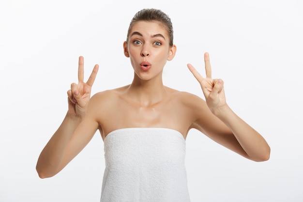 Крупный план красивой молодой женщины с банным полотенцем, покрывающим грудь на сером, показывает знак двух пальцев