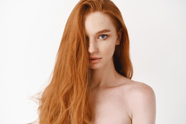 Closeup of beautiful young woman with long healthy red hair looking at camera Pale female redhead model gazing sensual white background
