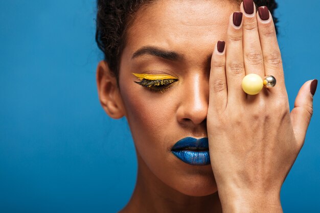 Closeup beauty relaxed mixed-race woman with fancy makeup posing on camera covering one eye with hand, isolated over blue wall