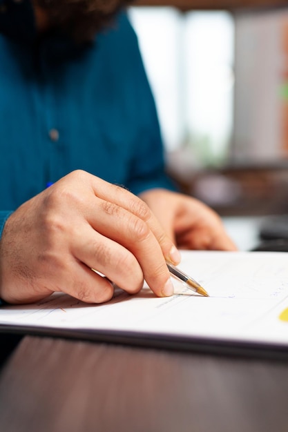 Closeup of businessman hand sitting at desk analyzing document with data chart on it working at management presentation writing business strategy. Entrepreneur brainstorming ideas in startup office