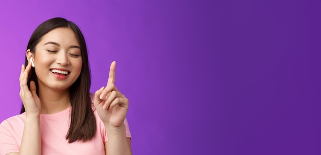 Free photo closeup cheerful glad attractive asian woman close eyes raising one finger waiting chorus smiling delighted touch wireless earphone make volume louder satisfied enjoy songs purple background