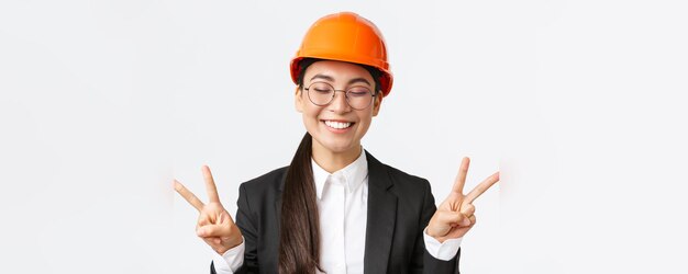 Closeup of cheerful successful female asian engineer construction architect in safety helmet and business suit showing peace signs and smiling kawaii standing white background