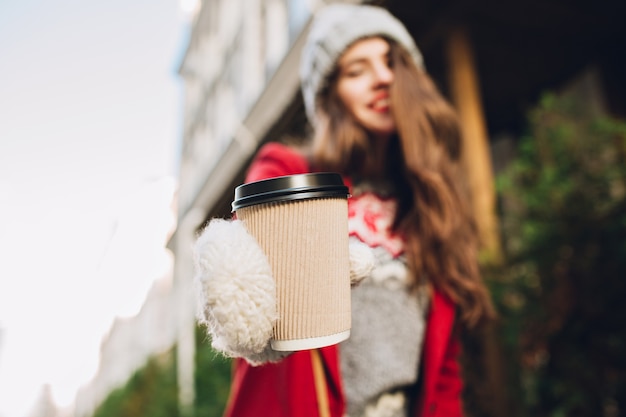 Кофе крупного плана, чтобы пойти растяжкой девушкой в белых перчатках на улице. Она носит красное пальто, у нее длинные волосы.