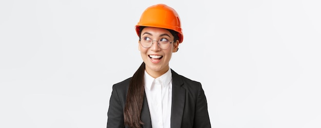 Крупный план творческой азиатской женщины-архитектора, имеющей в виду интересную идею инженера-строителя в деловом костюме и защитном шлеме, смотрящей влево с довольной улыбкой на белом фоне