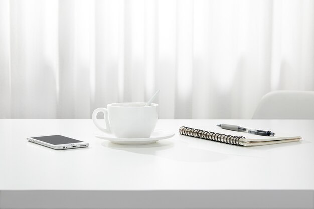Крупным планом чашка кофе, блокнот, ручка и смартфон на белом столе в помещении