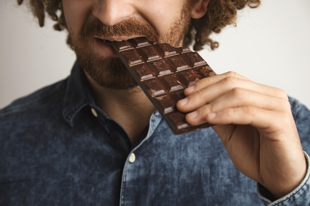 Primo piano capelli ricci felice uomo barbuto con pelle sana morde organico appena sfornato barretta di cioccolato con il lato della bocca, messa a fuoco ravvicinata sulla bocca