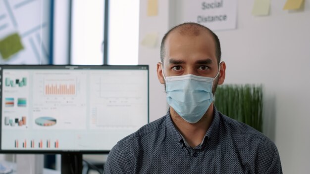 新しい通常の会社のオフィスでchiarに立っている間カメラを見て保護医療フェイスマスクを身に着けている従業員のクローズアップ。 covid19感染を回避するために社会的距離を尊重する労働者