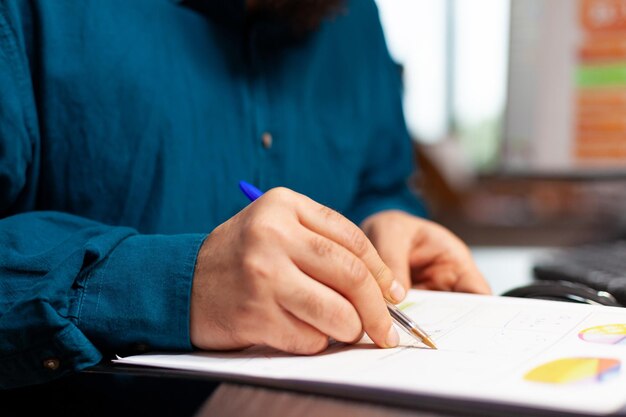 Closeup of entrepreneur hand sitting at table analyzing document with company plan on it working at management presentation writing business strategy. Businessman brainstorming ideas in startup office