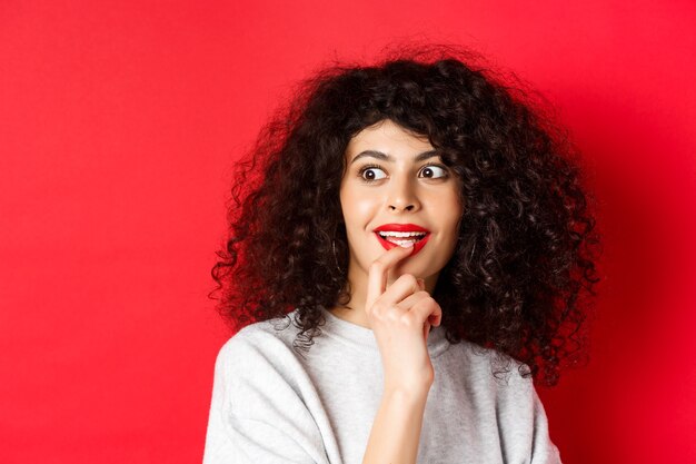 Closeup of excited curly woman touching lips and looking aside with interest checking out cool promo...