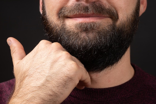 Closeup of face young man touches neat trimmed black thick beard with hand after skin and hair care ...