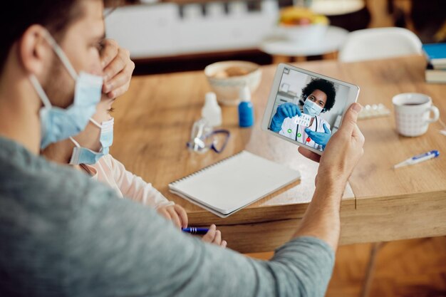 Крупный план отца и дочери, использующих сенсорную панель во время видеозвонка со своим семейным врачом из дома