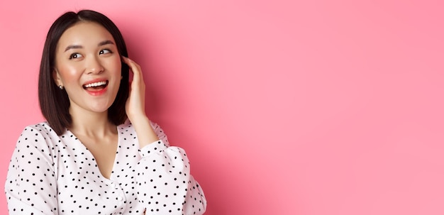 Free photo closeup of flirty asian woman sighing looking dreamy left and smiling standing over pink background