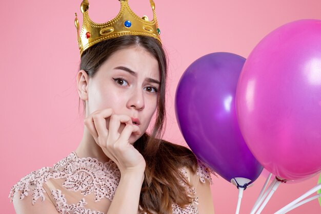 Closeup front view confused party girl with crown holding balloons putting hand