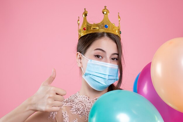 Closeup front view cute party girl with crown holding balloons making thumb up sign